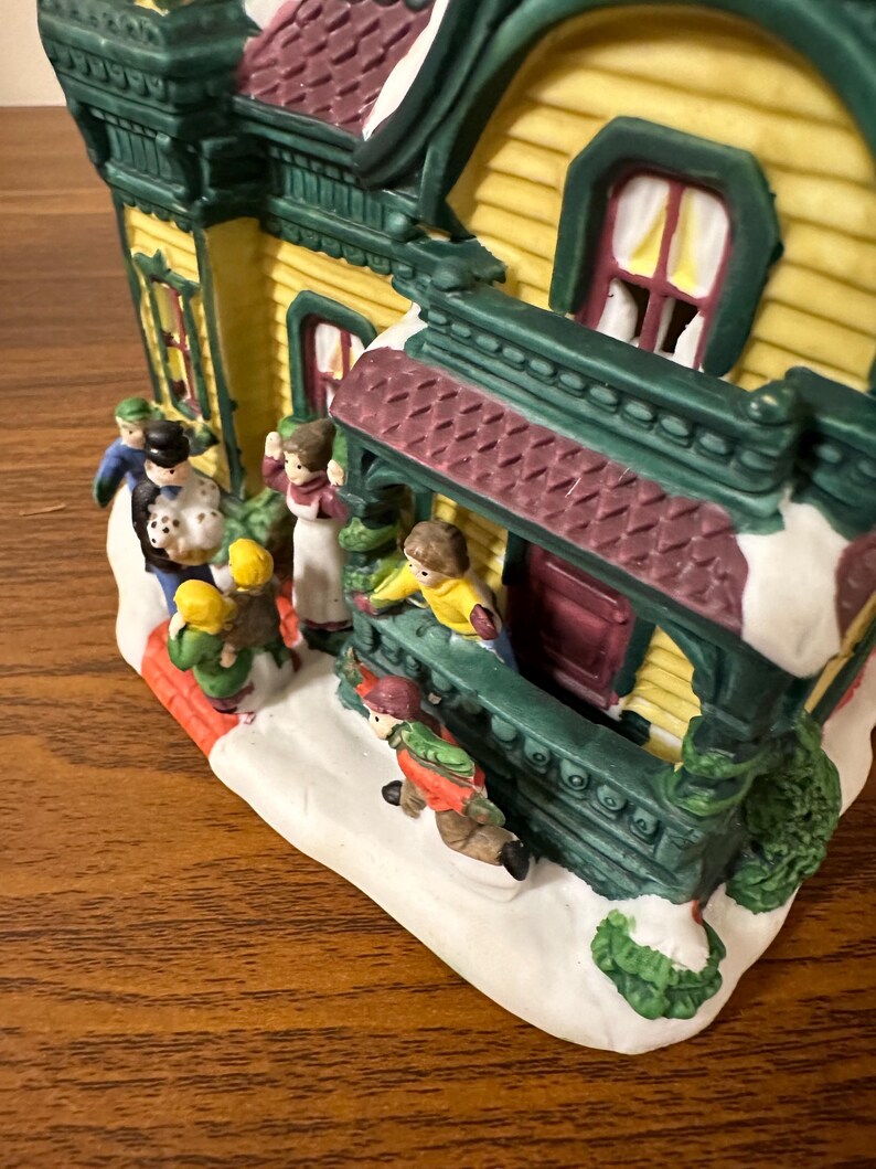 May include: A ceramic Christmas village house with a yellow facade, green trim, and a red roof. The house has a porch with a burgundy railing and a green door. There are small figures of people in front of the house, including a family of four and a couple.