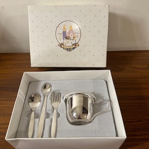 Puede incluir: Un juego de taza y cubiertos para bebé de plata en una caja blanca con una ilustración de conejo. La caja tiene un fondo blanco con pequeños puntos morados y las palabras "Briar Rose" en una pancarta morada.