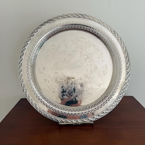 Puede incluir: Una bandeja redonda plateada con un diseño floral en relieve en el centro. La bandeja tiene un borde decorativo con un patrón repetitivo y un borde calado. Se muestra sobre una superficie de madera oscura.