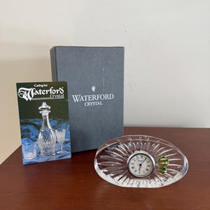 Puede incluir: Un reloj ovalado de cristal Waterford con esfera plateada y un trébol verde y dorado. El reloj está sobre una superficie de madera, junto a una caja gris y un folleto.