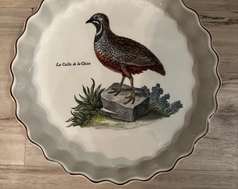 May include: A white ceramic baking dish with a scalloped rim and a brown and white illustration of a quail on a rock with the text "La Caille de la Chine" printed on the dish.
