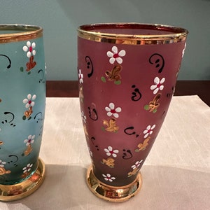 K&ouml;nnte beinhalten: Zwei Glasvasen mit goldfarbenem Rand und handbemaltem Blumendesign. Eine Vase ist hellblau mit gr&uuml;nen Akzenten und die andere ist dunkelrosa mit braunen Akzenten.