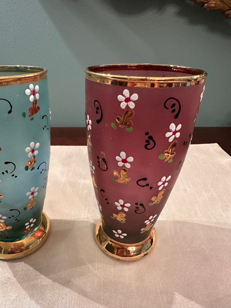 K&ouml;nnte beinhalten: Zwei Glasvasen mit goldfarbenem Rand und handbemaltem Blumendesign. Eine Vase ist hellblau mit gr&uuml;nen Akzenten und die andere ist dunkelrosa mit braunen Akzenten.