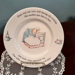 May include: A white ceramic plate featuring a blue and white illustration of Peter Rabbit in bed. The plate includes a quote from Beatrix Potter's "Peter Rabbit": "Peter was not very well during the evening. His mother put him to bed, and made some camomile tea: 'One table-spoon to be taken - bed-time.'"