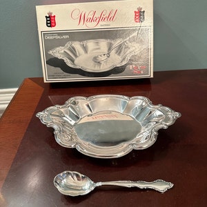 May include: A silver serving set with a small bowl and a spoon. The bowl has a scalloped edge and the spoon has a decorative handle. The set is labeled "Wakefield Pattern" and "DeepSilver".