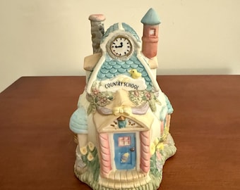 May include: A ceramic Country School figurine with a clock tower, blue roof, and a pink door. The figurine has a whimsical design with floral and mushroom accents. The text "COUNTRY SCHOOL" is visible on the front.