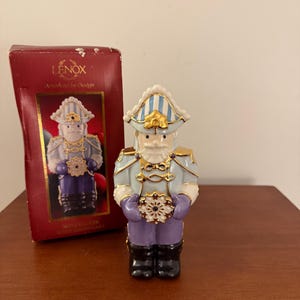 May include: A Lenox Nutcracker figurine with a snowflake design. The figurine is dressed in a light blue and purple uniform with gold accents. The box behind the figurine is red with the Lenox logo and the words "American by Design".