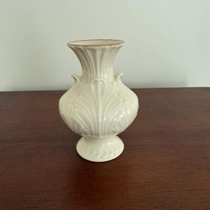 May include: Cream-coloured ceramic vase with a gold rim. The vase has a bulbous body with a detailed leaf pattern and two small handles. The base is round and also features a leaf pattern. A decorative piece for the home.