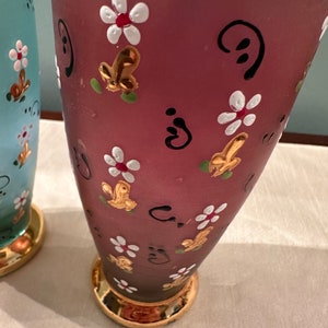 K&ouml;nnte beinhalten: Zwei Glasvasen mit goldenen R&auml;ndern und handbemalten Blumendesigns. Eine Vase ist hellblau mit wei&szlig;en Blumen und die andere ist tiefrosa mit wei&szlig;en Blumen und goldenen Akzenten.