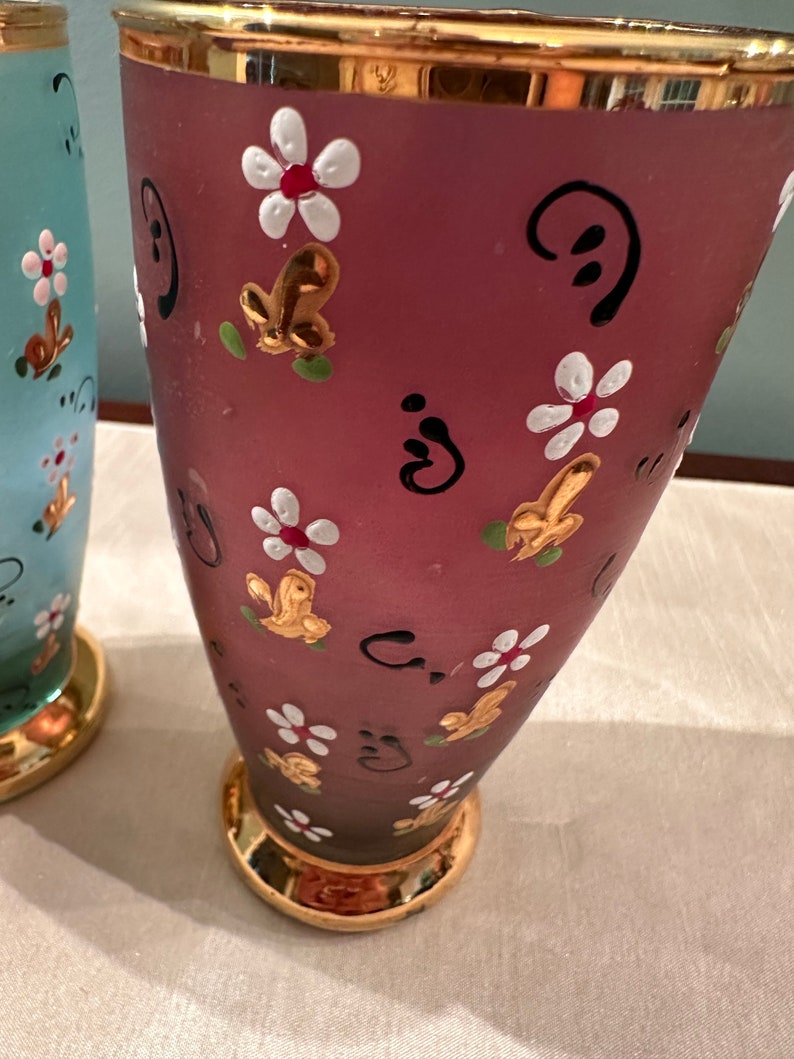 K&ouml;nnte beinhalten: Zwei Glasvasen mit goldenen R&auml;ndern und handbemalten Blumendesigns. Eine Vase ist hellblau mit wei&szlig;en Blumen und die andere ist tiefrosa mit wei&szlig;en Blumen und goldenen Akzenten.