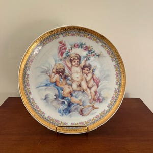 May include: A decorative porcelain plate featuring a classical scene with three cherubs amidst clouds. The plate has a gold rim with floral and pink accents. A lyre is also visible.