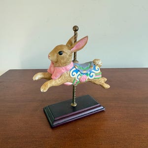 May include: A brown carousel rabbit figurine with pink ears and a pink collar. The rabbit has a blue and green saddle with floral accents and is mounted on a brass pole and a black base. The figurine is a decorative item.