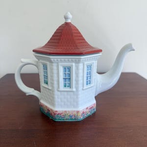 Op de afbeelding: Witte keramische theepot in de vorm van een huis met een rood dak. De theepot heeft een handvat, een tuit en een deksel. Het huisontwerp omvat ramen met blauwe ruiten en een bloemenrand aan de basis.