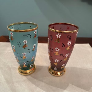 K&ouml;nnte beinhalten: Zwei handbemalte Glasbecher mit goldenen R&auml;ndern. Einer ist blau mit wei&szlig;en Blumen und der andere ist burgunderrot mit wei&szlig;en Blumen.