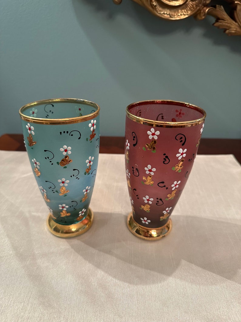 K&ouml;nnte beinhalten: Zwei handbemalte Glasbecher mit goldenen R&auml;ndern. Einer ist blau mit wei&szlig;en Blumen und der andere ist burgunderrot mit wei&szlig;en Blumen.