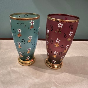 K&ouml;nnte beinhalten: Zwei Glasvasen mit goldenen R&auml;ndern und Blumenmustern. Eine Vase ist hellblau mit wei&szlig;en Blumen und die andere ist dunkelviolett mit wei&szlig;en Blumen.