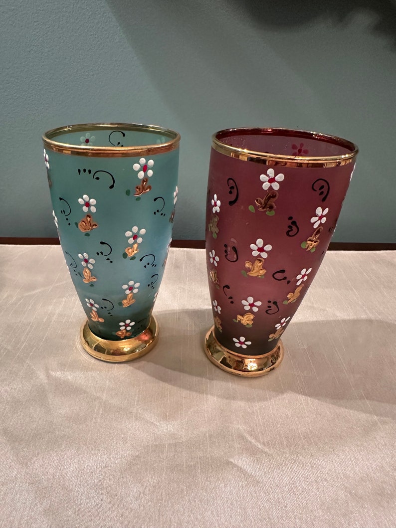 K&ouml;nnte beinhalten: Zwei Glasvasen mit goldenen R&auml;ndern und Blumenmustern. Eine Vase ist hellblau mit wei&szlig;en Blumen und die andere ist dunkelviolett mit wei&szlig;en Blumen.