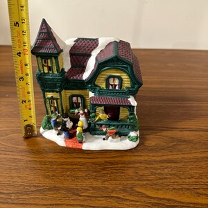 May include: A ceramic Christmas village house with a green and yellow exterior, a brown roof with white snow, and a red brick walkway.  There are small figurines of people in front of the house.