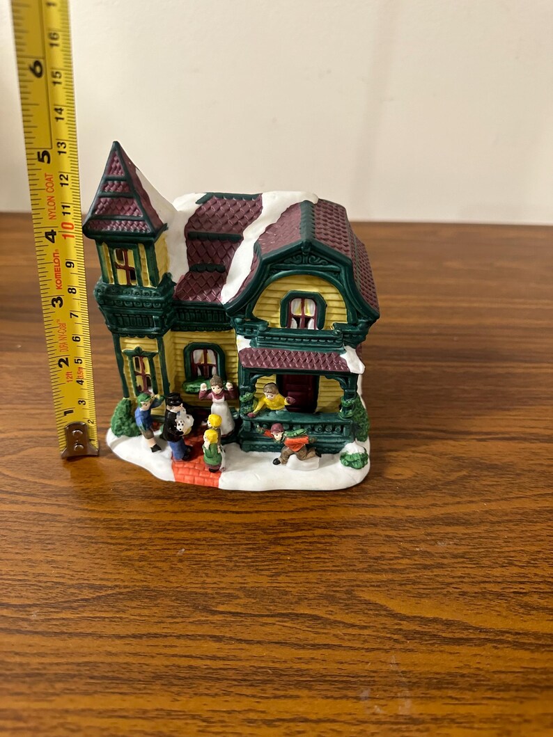 May include: A ceramic Christmas village house with a green and yellow exterior, a brown roof with white snow, and a red brick walkway.  There are small figurines of people in front of the house.