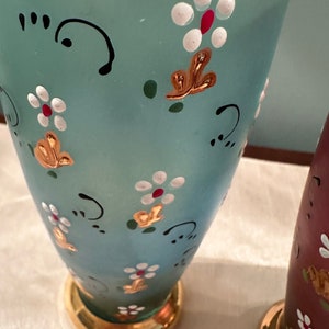 K&ouml;nnte beinhalten: Zwei Glasvasen mit goldenen Verzierungen und bemalten Blumendesigns. Eine Vase ist hellblau mit wei&szlig;en und roten Blumen, die andere ist dunkelrot mit wei&szlig;en Blumen.