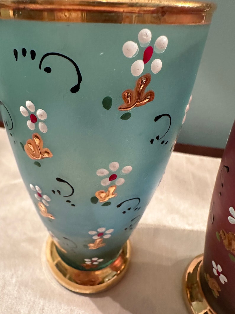 K&ouml;nnte beinhalten: Zwei Glasvasen mit goldenen Verzierungen und bemalten Blumendesigns. Eine Vase ist hellblau mit wei&szlig;en und roten Blumen, die andere ist dunkelrot mit wei&szlig;en Blumen.