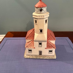 Könnte beinhalten: Eine Keramik-Leuchtturm-Figur mit rotem Dach, weißen Wänden und einem grau-weiß gestreiften Oberteil. Der Leuchtturm hat eine kleine Tür und Fenster.