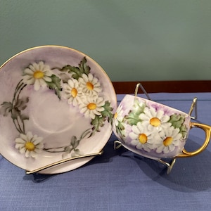 May include: A vintage teacup and saucer set with a delicate floral design. The cup and saucer are painted with white daisies on a lavender background. The edges of the cup and saucer are trimmed with gold.