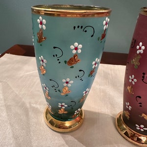 K&ouml;nnte beinhalten: Zwei antike Glasvasen mit goldenen Verzierungen und Blumendesigns. Die Vase links ist hellblau mit wei&szlig;en Blumen und goldenen Akzenten. Die Vase rechts ist in einem dunkleren Blauton mit wei&szlig;en Blumen und goldenen Akzenten.