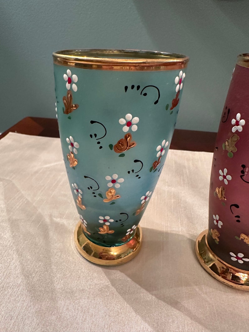 K&ouml;nnte beinhalten: Zwei antike Glasvasen mit goldenen Verzierungen und Blumendesigns. Die Vase links ist hellblau mit wei&szlig;en Blumen und goldenen Akzenten. Die Vase rechts ist in einem dunkleren Blauton mit wei&szlig;en Blumen und goldenen Akzenten.