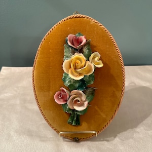 May include: A vintage oval-shaped wall plaque with a velvet backing in a mustard yellow color. The plaque features a 3D floral design with three porcelain roses in shades of pink, yellow, and white.