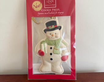 May include: A Lenox snowman cookie press ornament in a red package. The snowman has a black top hat, green scarf, red mittens, and holds a green and white gift. The package reads "New!" and "Cookie Press."