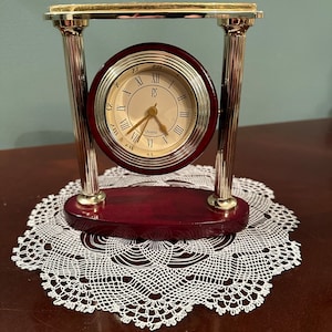 May include: A gold-toned clock with Roman numeral markings on a white face. The clock is on a wooden base with gold accents and is supported by two gold columns.