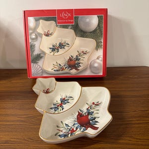 Può includere: Un piatto da portata in ceramica bianca a forma di albero di Natale con un bordo dorato. Il piatto è diviso in tre sezioni e presenta un design con un cardinale e bacche di agrifoglio. Il piatto è nella sua confezione originale, che presenta il logo Lenox e il testo "American by Design".