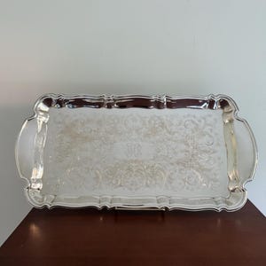 May include: A rectangular silver serving tray with detailed floral and scrollwork patterns. The tray has curved handles and a decorative edge. The centre of the tray features an engraved monogram. It is displayed on a dark wooden surface.