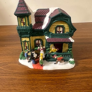 May include: A ceramic Christmas village house with a yellow facade, green trim, and a red roof. The house has a porch with a red brick walkway and a snow-covered roof. There are several small figures of people in front of the house, including a man in a black coat, a woman in a red dress, and a child in a blue coat.