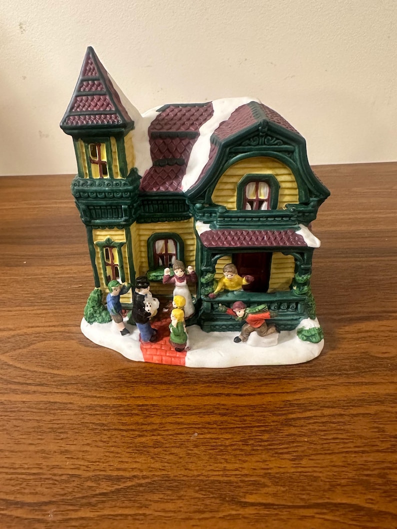 May include: A ceramic Christmas village house with a yellow facade, green trim, and a red roof. The house has a porch with a red brick walkway and a snow-covered roof. There are several small figures of people in front of the house, including a man in a black coat, a woman in a red dress, and a child in a blue coat.