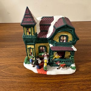 May include: A ceramic Christmas village house with a green roof, red trim, and yellow siding. The house has a snow-covered roof and a porch with people standing on it. There are also people walking on a brick path in front of the house.