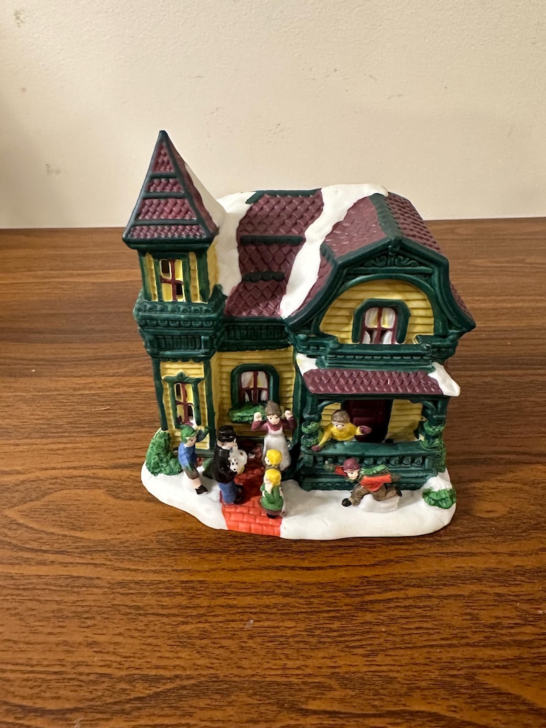 May include: A ceramic Christmas village house with a green roof, red trim, and yellow siding. The house has a snow-covered roof and a porch with people standing on it. There are also people walking on a brick path in front of the house.