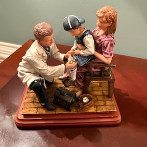 Puede incluir: Una figura que representa a un médico examinando la pierna de un niño, sostenido por una mujer. El médico lleva una bata blanca, y el niño una gorra y chaleco azules. También hay un guante de béisbol y un pequeño maletín negro.