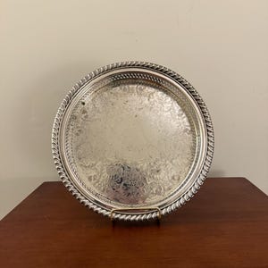Pode incluir: Uma bandeja redonda, prateada, com um design floral ornamentado em relevo no centro. A bandeja tem uma borda decorativa em forma de corda e é exibida em um suporte dourado. A bandeja está sobre uma superfície de madeira marrom escura.