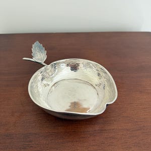 Może przedstawiać: Srebrna miska w kształcie jabłka z teksturowaną powierzchnią i uchwytem w kształcie liścia. Misa wykonana jest z błyszczącego metalu i stoi na ciemnej drewnianej powierzchni. Misa jest pusta.