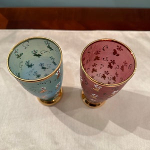K&ouml;nnte beinhalten: Zwei gl&auml;serne Becher mit goldenen R&auml;ndern und Blumenmustern. Ein Becher ist hellblau mit gr&uuml;nen und wei&szlig;en Blumen, der andere ist rosa mit braunen und wei&szlig;en Blumen.