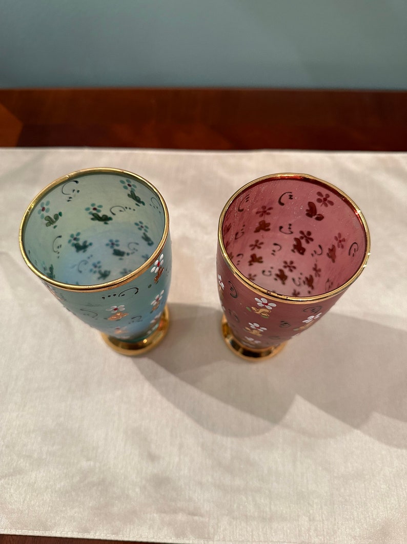 K&ouml;nnte beinhalten: Zwei gl&auml;serne Becher mit goldenen R&auml;ndern und Blumenmustern. Ein Becher ist hellblau mit gr&uuml;nen und wei&szlig;en Blumen, der andere ist rosa mit braunen und wei&szlig;en Blumen.