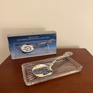 May include: Silver-plated cranberry server with a glass liner. The server features an ornate handle and a slotted spoon. The glass liner is rectangular with a sunburst design. The box reads "Silver Plated Cranberry Server with Glass Liner."