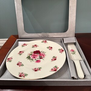 Könnte beinhalten: Ein weißes Kuchenplatten- und Servierset mit einem Blumendesign mit rosa und roten Rosen. Das Set befindet sich in seiner Originalverpackung, auf der "Formalities™ by Baum Bros." und "Cake Plate & Server" steht.