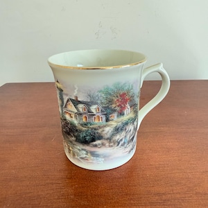 Può includere: Tazza in ceramica color crema con manico e bordo dorato. La tazza presenta un'illustrazione panoramica di una casa e di un faro su una scogliera, con un albero autunnale colorato. La tazza è di dimensioni standard per caffè o tè.