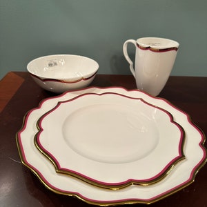 May include: A set of white ceramic dinnerware with a scalloped edge and a gold and burgundy trim. The set includes a bowl, a plate, and a mug.