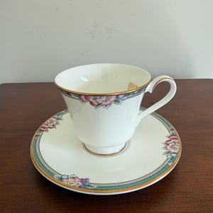 Op de afbeelding: Een witte keramische mok met een zwart handvat en de tekst "But First, Coffee" in een zwarte, handgeschreven lettertype. De mok staat op een wit oppervlak.