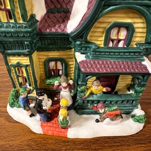 May include: A ceramic Christmas village house with a yellow facade, green trim, and a red roof. The house has a porch with a railing and a white snow-covered roof. There are several small figurines of people in front of the house.