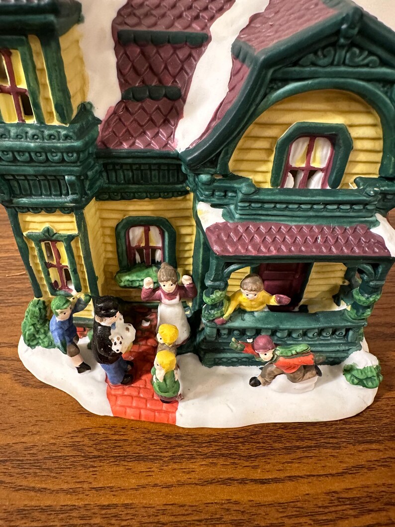 May include: A ceramic Christmas village house with a yellow facade, green trim, and a red roof. The house has a porch with a railing and a white snow-covered roof. There are several small figurines of people in front of the house.
